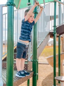 A young boy with light skin is on a silver and green playground waiting to hang from monkey bars. He is wearing a blue and black striped shirt with monster trucks, black shorts, and black Stride Rite shoes.
